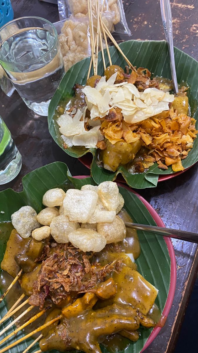 Dua porsi sate padang berisi sate dan lontong yang dilumuri dengan saus sate padang yang kental dan disajikan di atas piring dan daun pisang. Porsi pertama dilengkapi dengan topping kerupuk kulit dan porsi kedua dilengkapi dengan keripik singkong